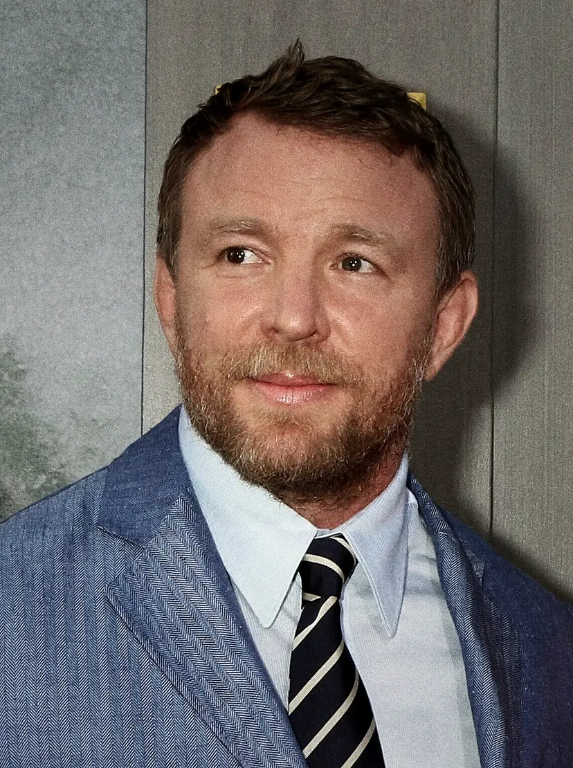 Guy Ritchie in a suit and open-necked shirt, smiling