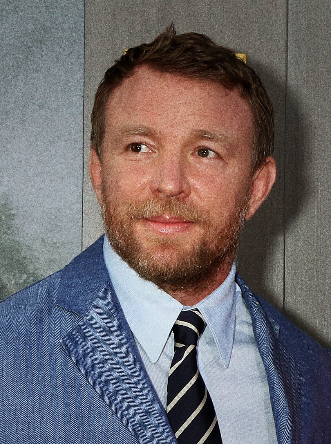 Guy Ritchie in a suit and open-necked shirt, smiling